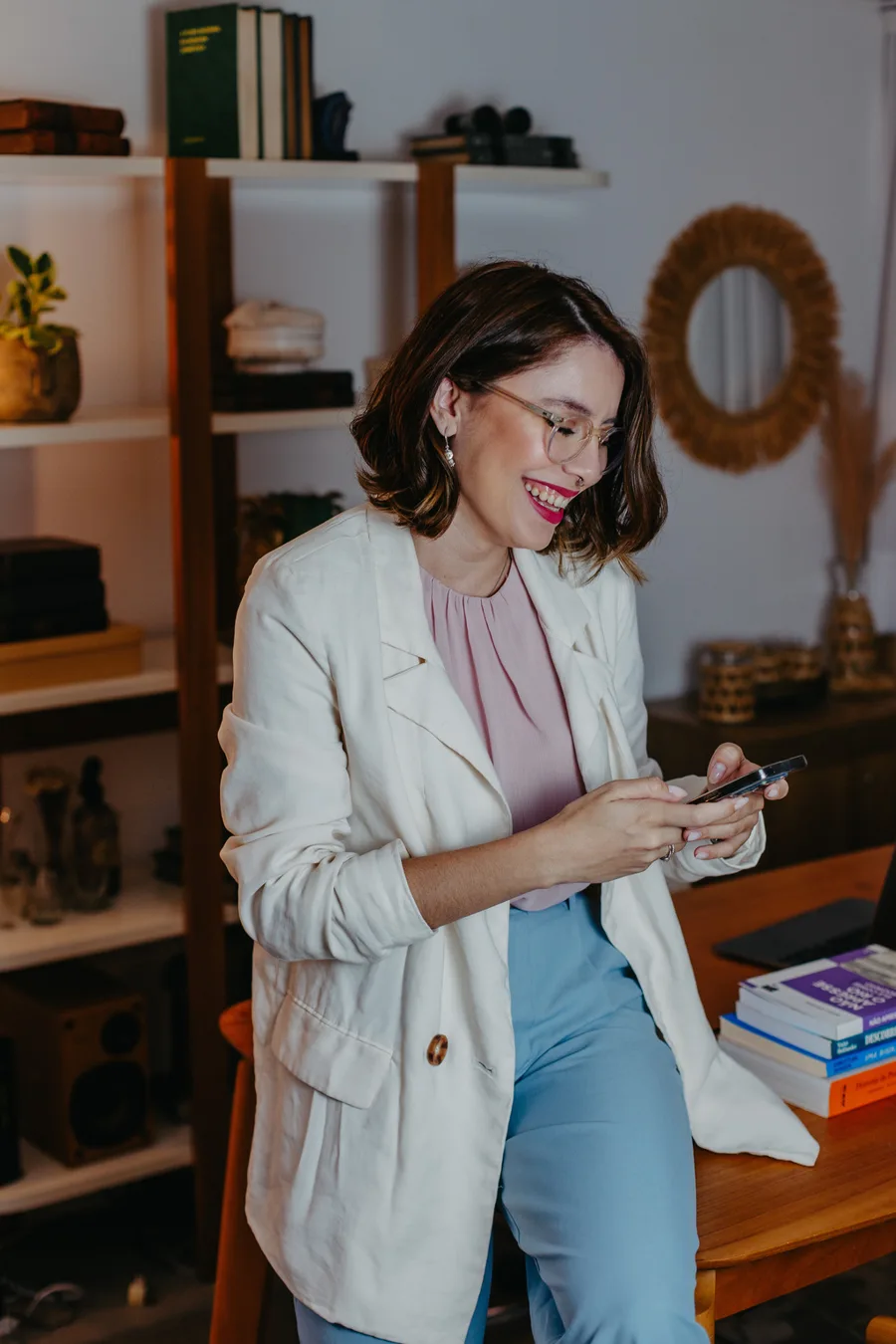 Layana Belyssa sorrindo, olhando para o celular, em uma sala com estante e livros